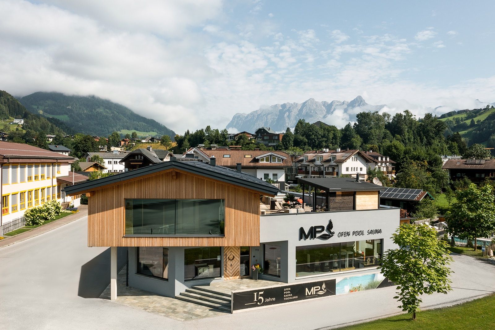 Ein modernes Gebäude mit Holzfassade und Glasfenstern steht in einem Dorf mit Häusern und Bergen im Hintergrund. Es hat ein Schild mit MP-Logo und Text.