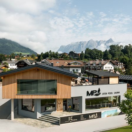 Ein modernes Gebäude mit Holzverkleidung und großen Fenstern, gekennzeichnet mit MP, mit Bergen und Häusern im Hintergrund.