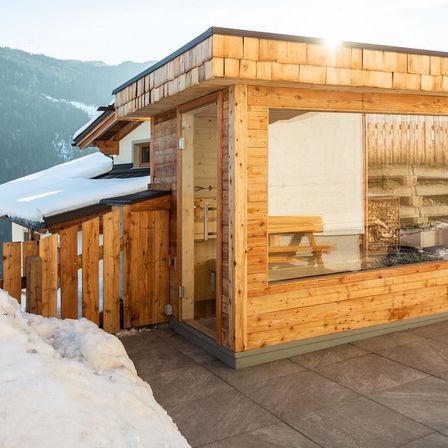 Ein Holzchalet mit Schnee auf dem Dach und einer Bank vor einer Glastür. Berge im Hintergrund.