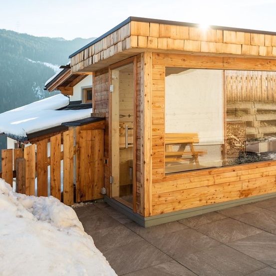 Ein Holzchalet mit Schnee auf dem Dach und einer Bank vor einer Glastür. Berge im Hintergrund.