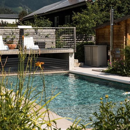 Ein ruhiger Außenpool mit klarem blauem Wasser, umgeben von üppigen Pflanzen und Blumen. Ein Holzhaus mit Solarpaneelen ist im Hintergrund.