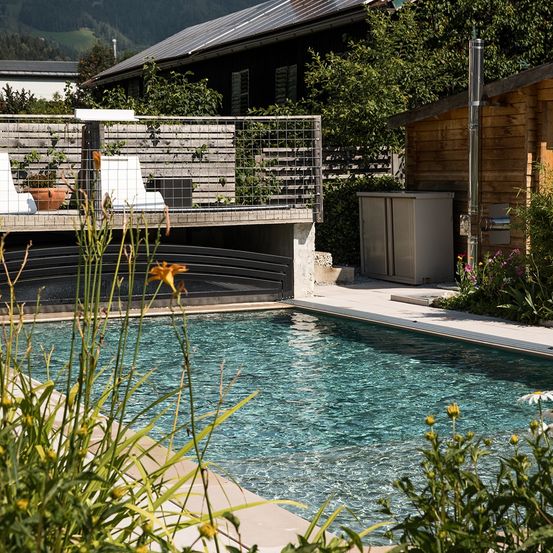 Ein ruhiger Außenpool mit klarem blauem Wasser, umgeben von üppigen Pflanzen und Blumen. Ein Holzhaus mit Solarpaneelen ist im Hintergrund.