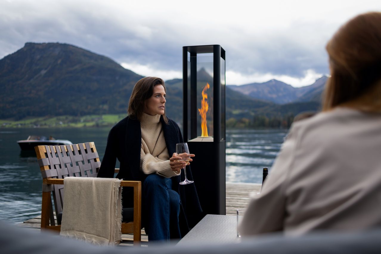 A woman holding a wine glass sits on a chair by a lake, with a fire pit in the background.