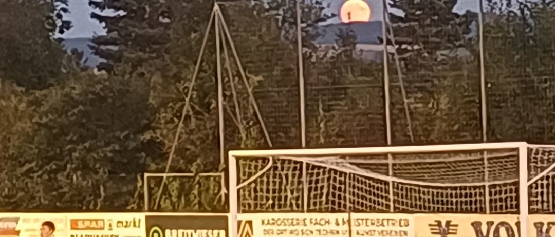 Ein Fußballspieler steht auf einem Feld unter einem untergehenden Mond, mit einem Netz und Werbebannern im Hintergrund.