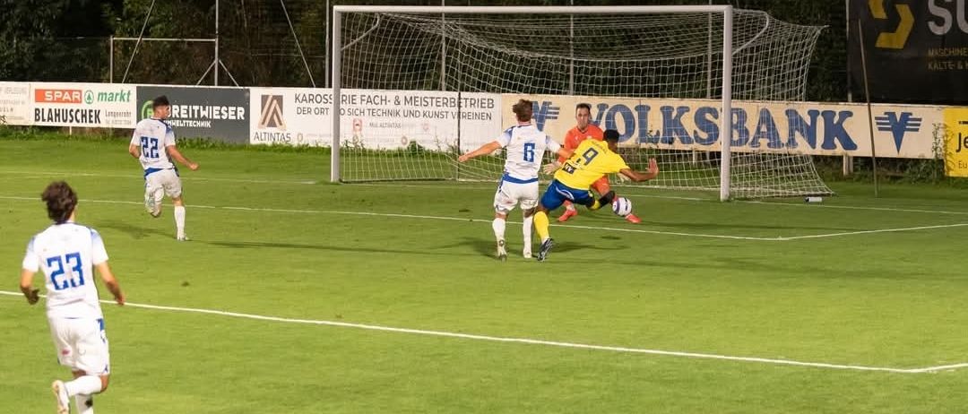 Ein Fußballspiel findet bei Nacht auf einem Feld statt. Spieler laufen, einer in blau-gelben Uniformen versucht zu treffen. Zwei Spieler in weißen Uniformen versuchen zu blocken. Hinter dem Tor befinden sich Plakate.