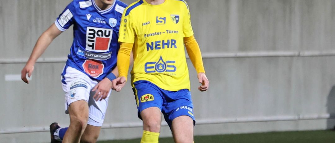 Zwei Fußballspieler, einer in Blau und Weiß, der andere in Gelb, laufen auf dem Feld. Sie jagen einem weißen Ball hinterher. Dahinter befindet sich eine Betonmauer mit leeren Sitzen.