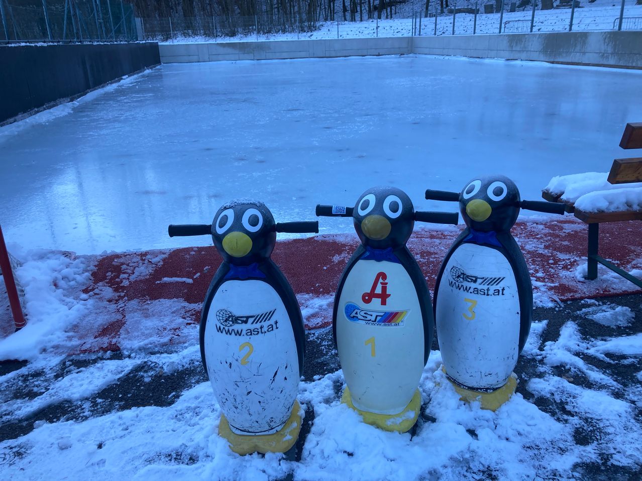 Drei Pinguinstatuen mit den Nummern 1, 2 und 3 stehen auf einer verschneiten Oberfläche, mit einer großen Eisbahn im Hintergrund.