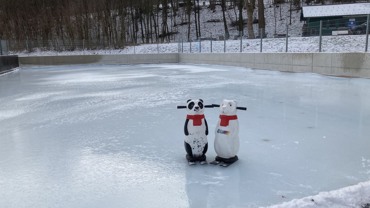 Zwei Schneemann-Statuen stehen auf einer Eisbahn. Sie tragen rote Schals und halten Skistöcke. Die Bahn ist von einer Betonbarriere umgeben.