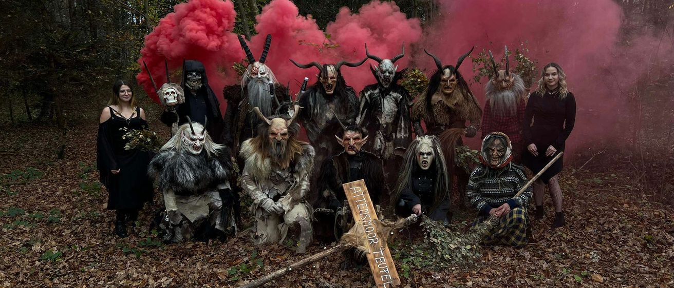 Gruppe von Menschen in gruseligen Kostümen posiert in einem Wald mit rotem Rauch im Hintergrund. Ein Holzschild ist im Vordergrund platziert.