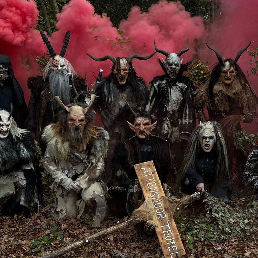 Gruppe von Menschen in gruseligen Kostümen posiert in einem Wald mit rotem Rauch im Hintergrund. Ein Holzschild ist im Vordergrund platziert.