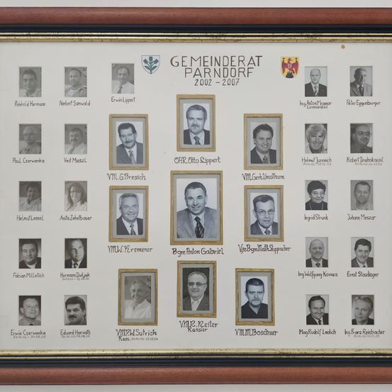 Ein gerahmtes Collage von 24 Porträts von Personen, mit Namen und Daten beschriftet, auf weißem Hintergrund mit goldener Einfassung. Die Collage trägt den Titel 'Gemeinderat Parndorf 2002-2007'.