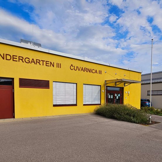 Ein leuchtend gelbes Gebäude mit den Worten Kindergarten III und Cuvarnica III darauf. Es hat eine rote Tür und drei Fenster mit weißen Rollläden.