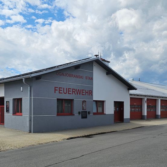 Ein Feuerwehrgebäude mit grauen Wänden, rotem Zierrat und dem Wort 'Feuerwehr' in Rot geschrieben. Das Gebäude hat mehrere Fenster und eine rote Tür.
