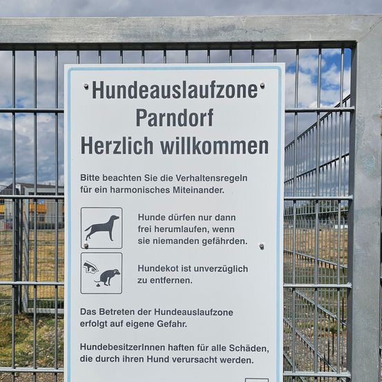 Ein Schild auf Deutsch am Eingang eines Hundeparks, das Besucher willkommen heißt und Regeln für Hundebesitzer bereitstellt. Das Schild enthält ein Bild eines Hundes und Anweisungen für das Hundeverhalten.