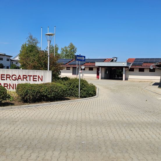 Eine gepflasterte Auffahrt führt zu einem Gebäude mit einem Schild, das Kindergarten steht. Solarmodule sind auf dem Dach installiert.