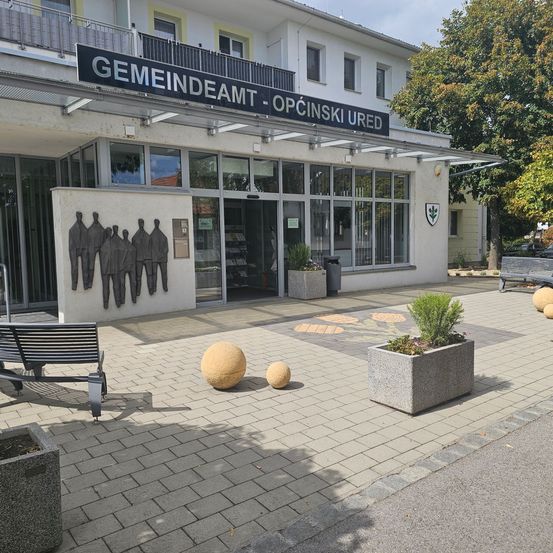 Der Eingang zu einem Gemeindeamt mit einem Schild, das 'Gemeindeamt - Opcinski Ured' besagt. Der Eingang wird von einer Wand mit Statuen und einer Bank flankiert.