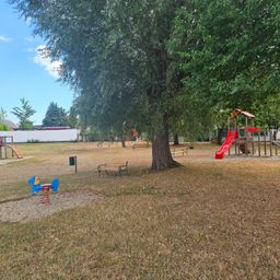 Ein Kinderspielplatz mit einer roten Rutsche, einem blauen Schaukelpferd und Bänken unter einem großen Baum. Der Bereich ist von Gras und Bäumen umgeben.