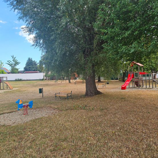 Ein Kinderspielplatz mit einer roten Rutsche, einem blauen Schaukelpferd und Bänken unter einem großen Baum. Der Bereich ist von Gras und Bäumen umgeben.