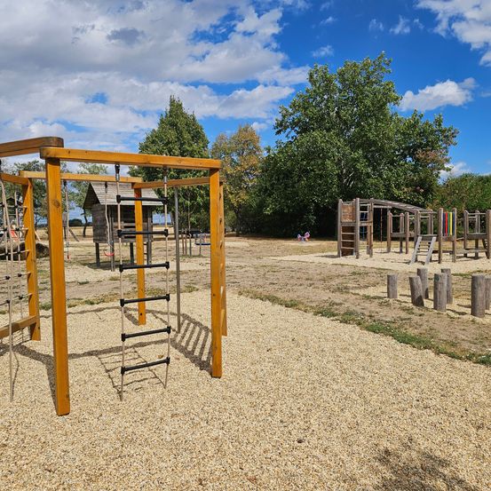 Ein Outdoor-Spielplatz mit Holzkonstruktionen, einer Leiter und einem Schaukelset unter einem blauen Himmel mit Wolken.