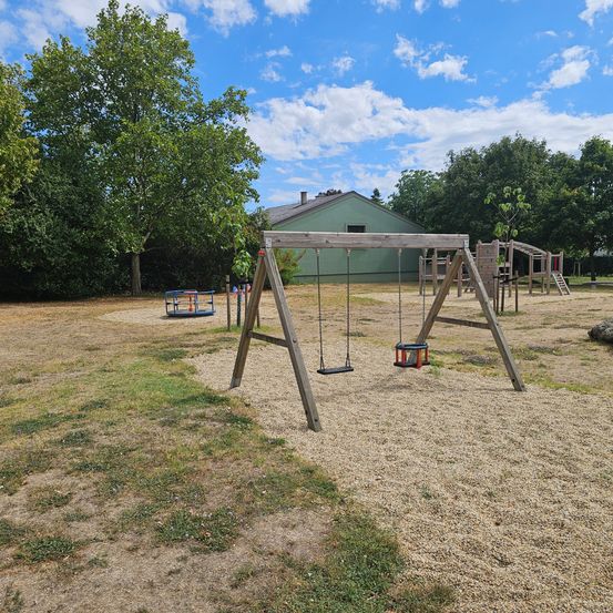 Ein Spielplatz mit Schaukel und Klettergerüst, umgeben von Bäumen und Gras unter blauem Himmel.