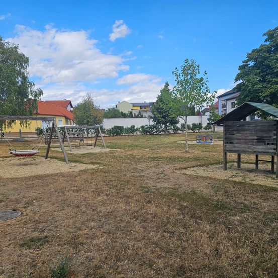 Ein Spielplatz mit Schaukeln und einem kleinen Holzhaus unter einem blauen Himmel mit verstreuten Wolken.