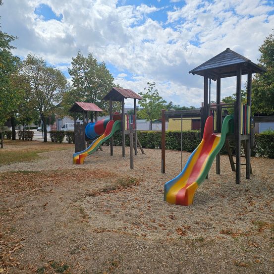 Zwei bunte Rutschen befinden sich in einem Spielplatz mit Bäumen und Büschen. Der Himmel ist blau mit einigen Wolken.