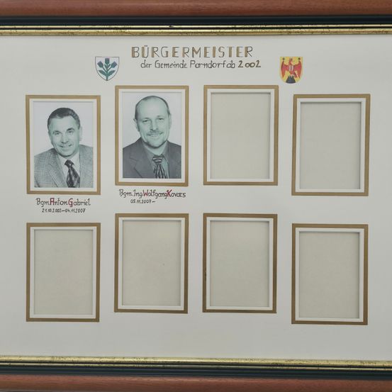 Ein gerahmter Rahmen mit goldenem Rand zeigt die Namen und Fotos ehemaliger Bürgermeister von Parndorf, Österreich. Nur zwei Fotos sind sichtbar, eines von Anton Gabriel und ein anderes von Ing. Walfang Kovats.