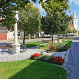 Ein gut gepflegter Park mit einem weißen Pfeiler, bunten Blumen und einer Kirche im Hintergrund an einem sonnigen Tag.