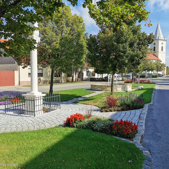 Ein gut gepflegter Park mit einem weißen Pfeiler, bunten Blumen und einer Kirche im Hintergrund an einem sonnigen Tag.