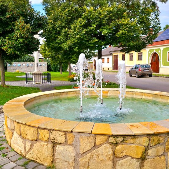 Ein runder Brunnen mit drei Wasserstrahlen steht in der Mitte eines grünen Parks. Dahinter befinden sich Bäume, ein Gebäude und ein geparktes Auto auf der Straße.
