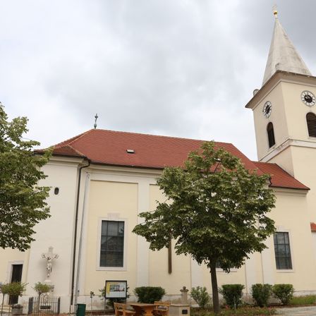 Eine weiße Kirche mit rotem Dach und einem Kirchturm mit einer Uhr. Sie hat eine Statue von Jesus Christus und ein Kreuz an der Wand. Bäume und Pflanzen umgeben die Kirche.