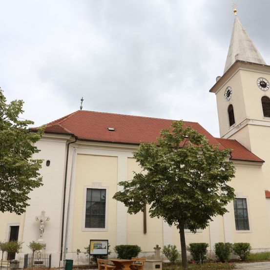 Eine weiße Kirche mit rotem Dach und einem Kirchturm mit einer Uhr. Sie hat eine Statue von Jesus Christus und ein Kreuz an der Wand. Bäume und Pflanzen umgeben die Kirche.