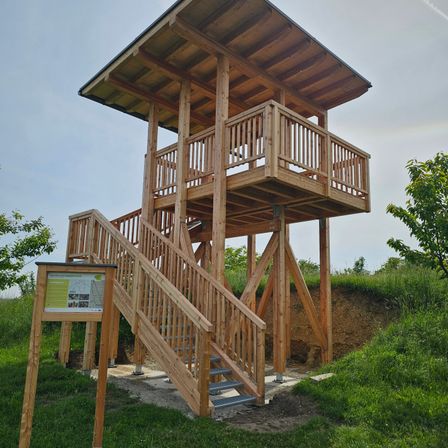 Ein hölzerner Aussichtsturm mit Treppen und Balkon, umgeben von grünem Gras und einem kleinen Schild.