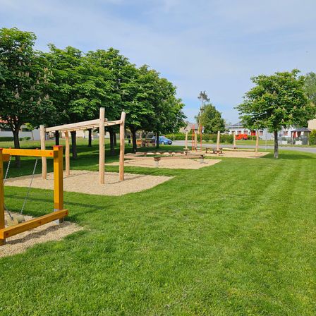 Spielplatz im Freien mit Holzstrukturen auf einem Rasenfeld, umgeben von Bäumen. Im Hintergrund sind ein Gebäude und ein geparktes Auto zu sehen.