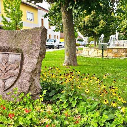 Ein Steinmonument mit einem gravierten Design steht inmitten eines farbenfrohen Gartens. Dahinter befindet sich eine Fontäne in einem grasbewachsenen Bereich. Bäume, Blumen und ein gelbes Gebäude mit geparkten Autos sind im Hintergrund zu sehen.