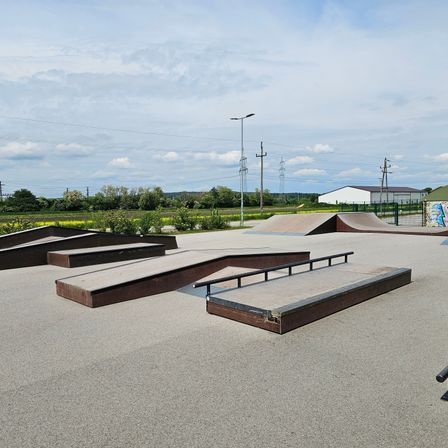 Ein Skatepark mit mehreren Rampen und einer ebenen Fläche. Die Rampen sind aus Beton und haben Metallhandläufe. Das Gelände ist von einem Zaun umgeben.