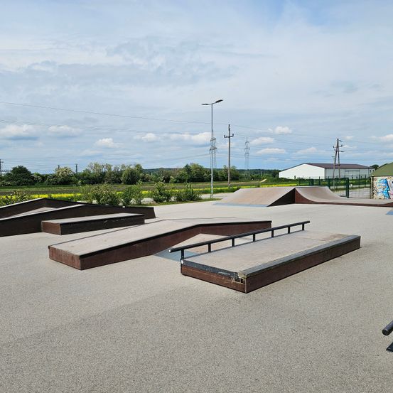 Ein Skatepark mit mehreren Rampen und einer ebenen Fläche. Die Rampen sind aus Beton und haben Metallhandläufe. Das Gelände ist von einem Zaun umgeben.