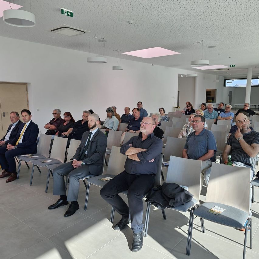 Eine Gruppe von Menschen in Anzügen und Brillen sitzen auf Stühlen in einem Konferenzraum mit grauen Stühlen, weißen Wänden und Lampen, die von der Decke hängen.