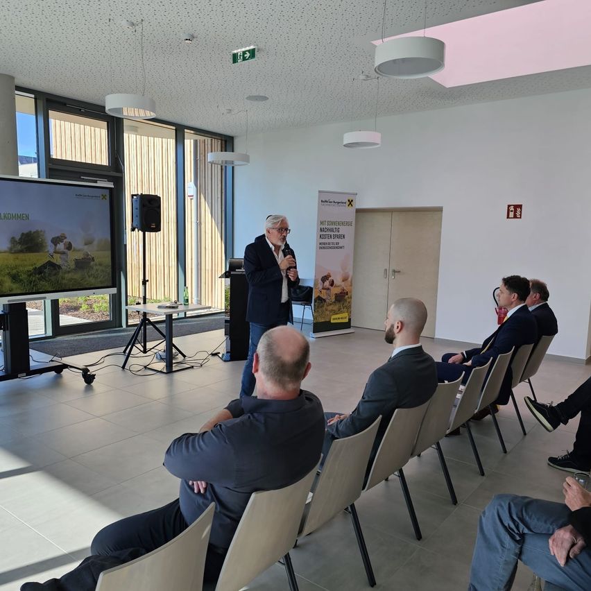Ein Mann hält eine Rede in einem Konferenzraum mit einer Projektionsleinwand hinter ihm. Mehrere Personen sitzen vor ihm auf Stühlen.
