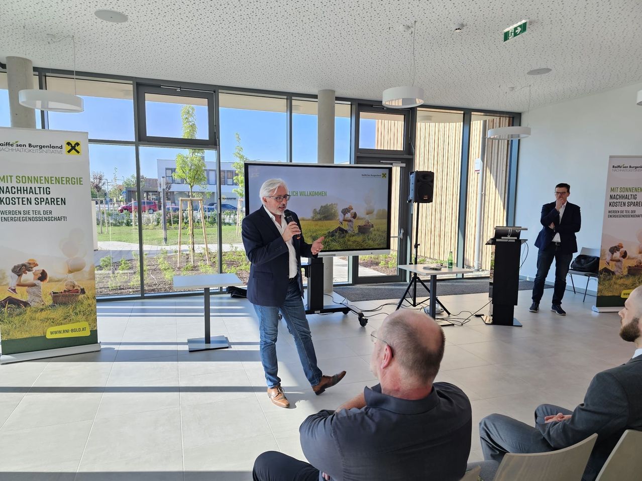 Ein Mann in Anzug und Brille hält in einem Konferenzraum eine Rede und hält ein Mikrofon in der Hand. Ein anderer Mann sitzt vor ihm.