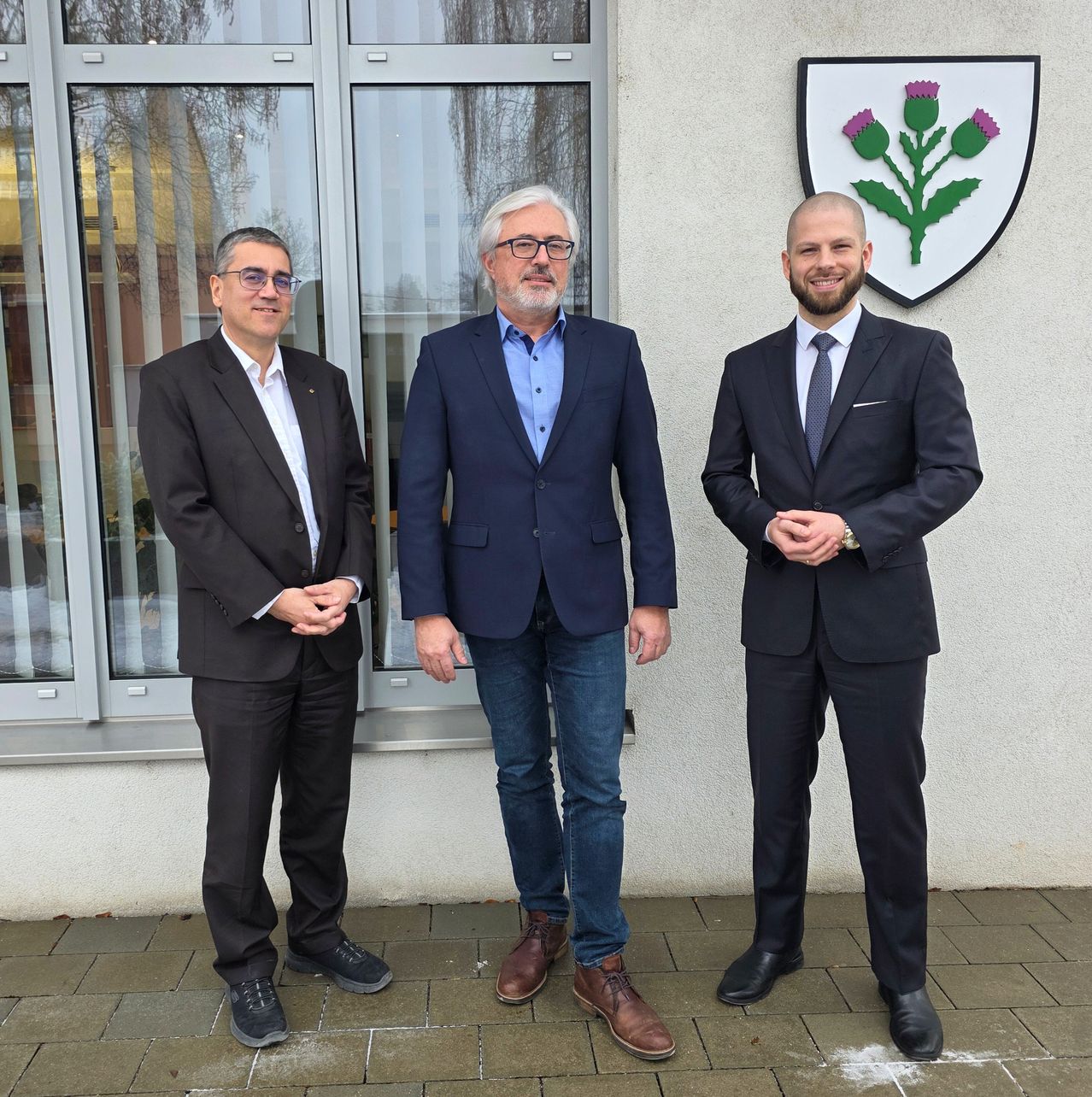 Drei Männer in Anzügen und Krawatten stehen vor einer weißen Wand mit einem Wappen, das einen Distel darstellt.