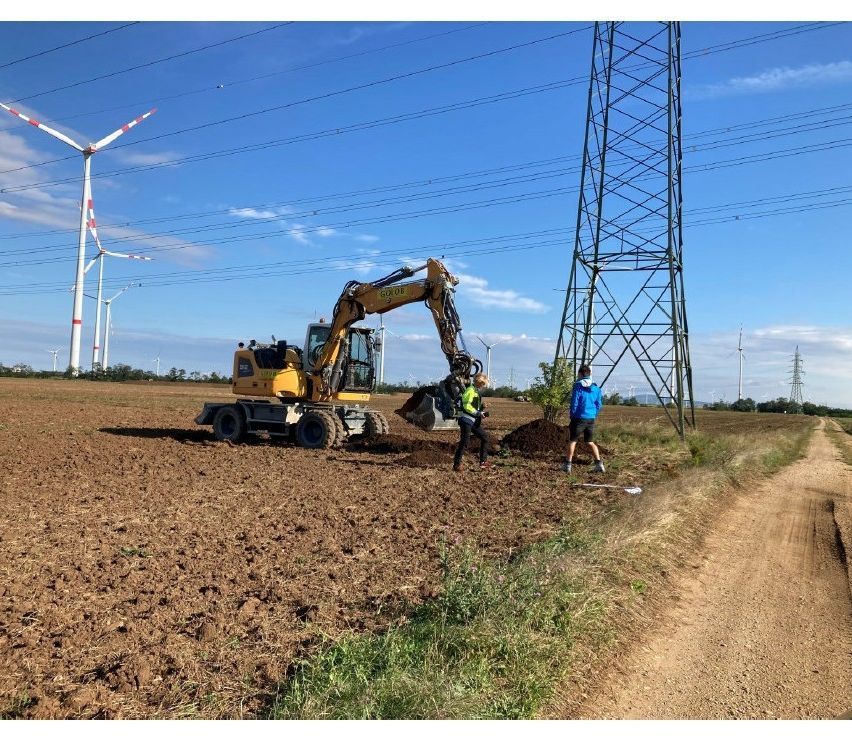 Zwei Personen auf einem Feld mit einem gelben Bagger, der ein Loch gräbt, einem Übertragungsturm und Windturbinen im Hintergrund.
