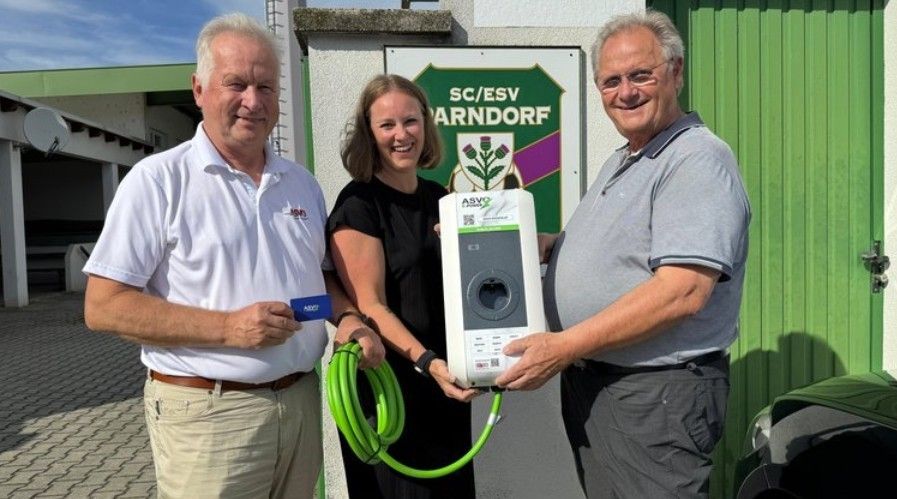 Drei Menschen stehen lächelnd vor einem Gebäude. Der Mann auf der linken Seite hält eine Karte und ein Kabel, während die Frau in der Mitte das andere Ende des Kabels hält. Der Mann auf der rechten Seite hält ein Gerät.