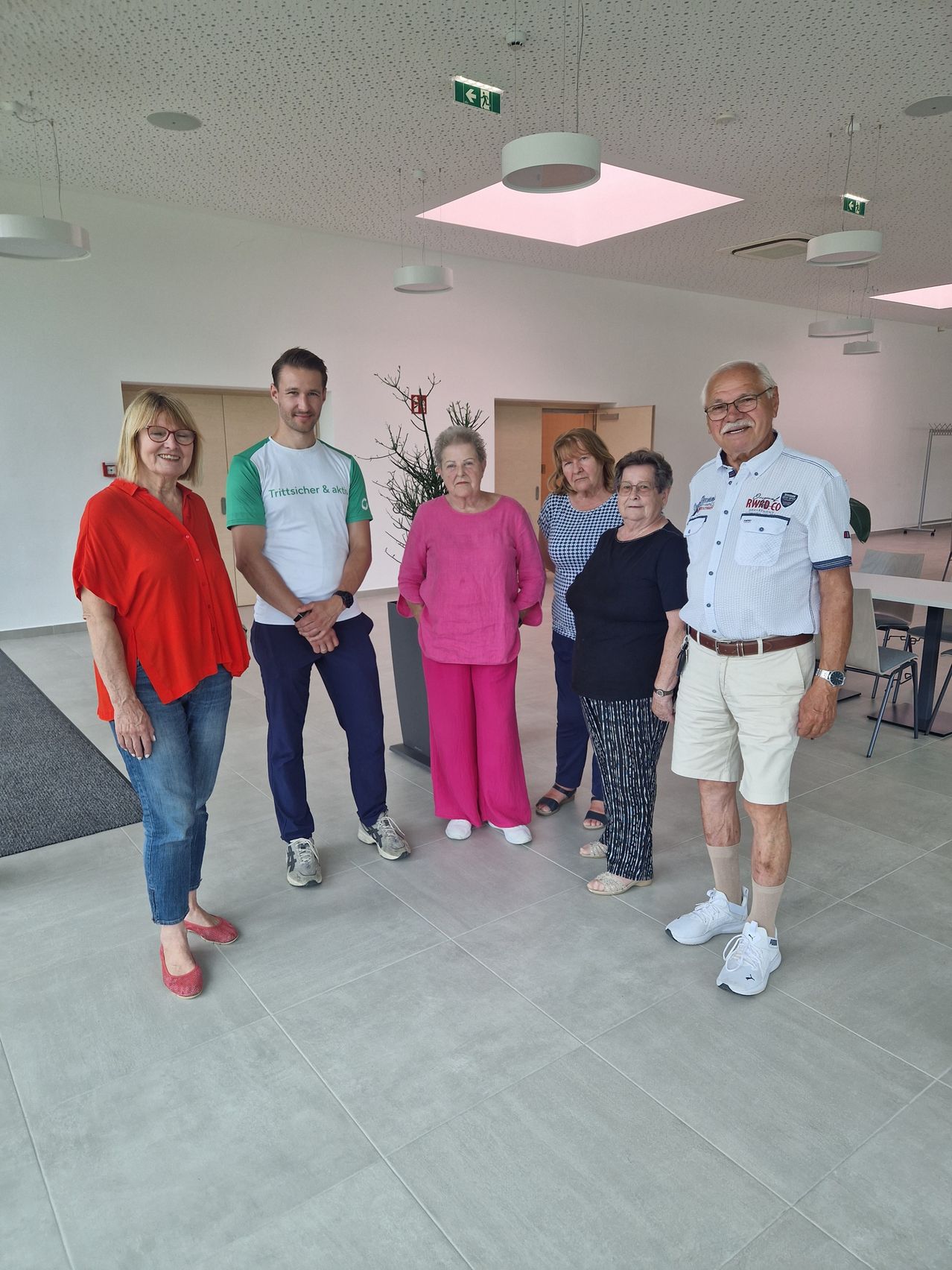 Sechs Personen, drei Frauen und drei Männer, stehen in einem Raum mit weißen Wänden und Deckenleuchten. Eine Frau in einem roten Blusenhemd steht neben einem Mann in einem weißen Shirt mit grünem Text. Dahinter steht eine Frau in rosa Hosen und einer Pflanze.