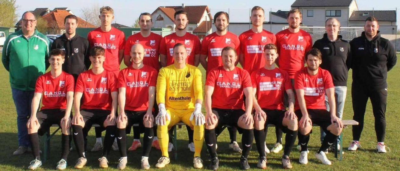 Eine Fußballmannschaft in roten Trikots posiert für ein Gruppenfoto auf einem Feld. Der Torwart in einem gelben Trikot steht davor. Gebäude und ein Zaun sind im Hintergrund.