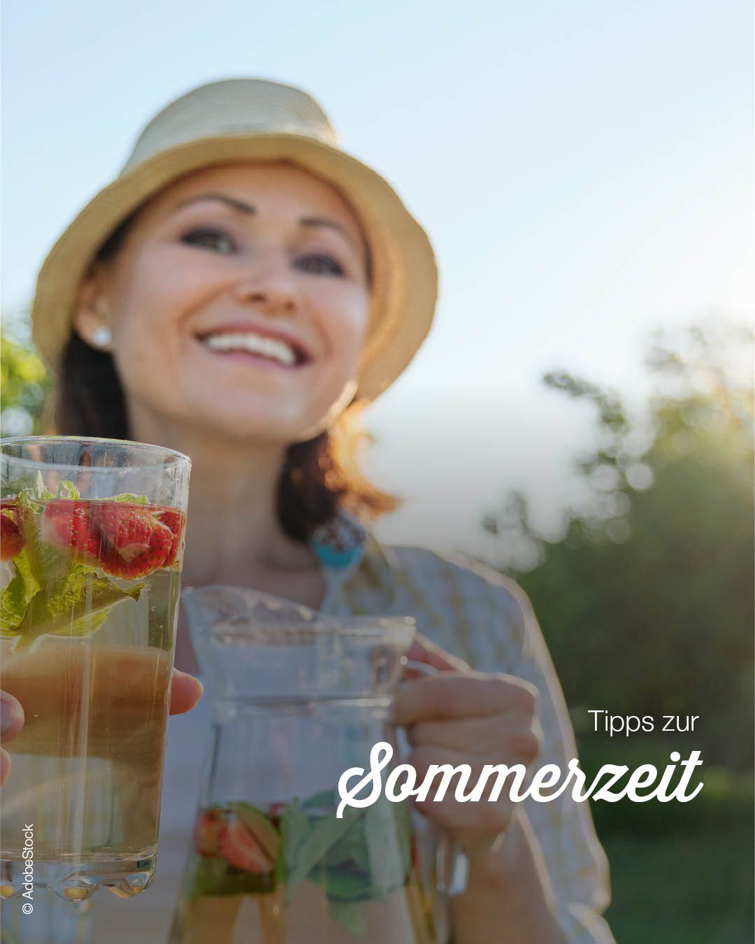 Eine Frau mit Hut lächelt und hält ein Glas Tee mit Erdbeeren und Minzblättern. Sie steht in einem Garten.