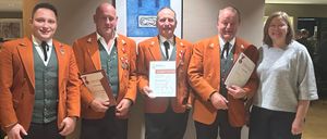 Fünf Personen in orangefarbenen Uniformen stehen lächelnd zusammen. Sie halten Urkunden in den Händen und eine Person hat eine gerahmte Urkunde. An der Wand hinter ihnen befindet sich ein blau gerahmtes Gemälde.