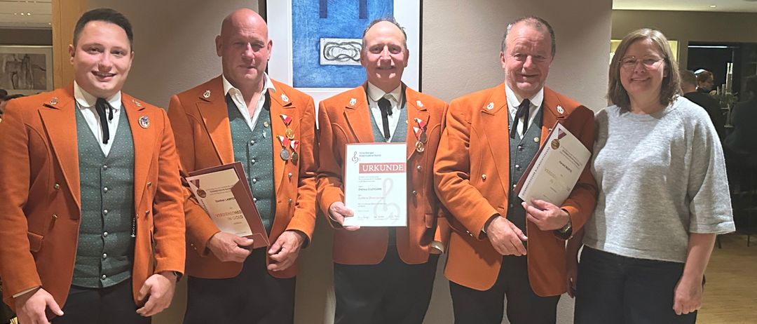 Fünf Personen in orangefarbenen Uniformen stehen lächelnd zusammen. Sie halten Urkunden in den Händen und eine Person hat eine gerahmte Urkunde. An der Wand hinter ihnen befindet sich ein blau gerahmtes Gemälde.