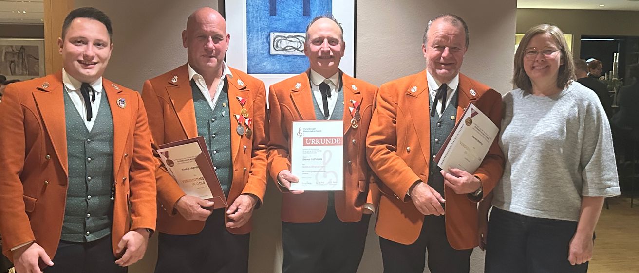 Fünf Personen in orangefarbenen Uniformen stehen lächelnd zusammen. Sie halten Urkunden in den Händen und eine Person hat eine gerahmte Urkunde. An der Wand hinter ihnen befindet sich ein blau gerahmtes Gemälde.