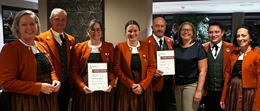 Eine Gruppe von Menschen in traditioneller Kleidung, darunter Frauen in orangefarbenen Jacken und braunen Röcken, stehen in einem Raum zusammen.